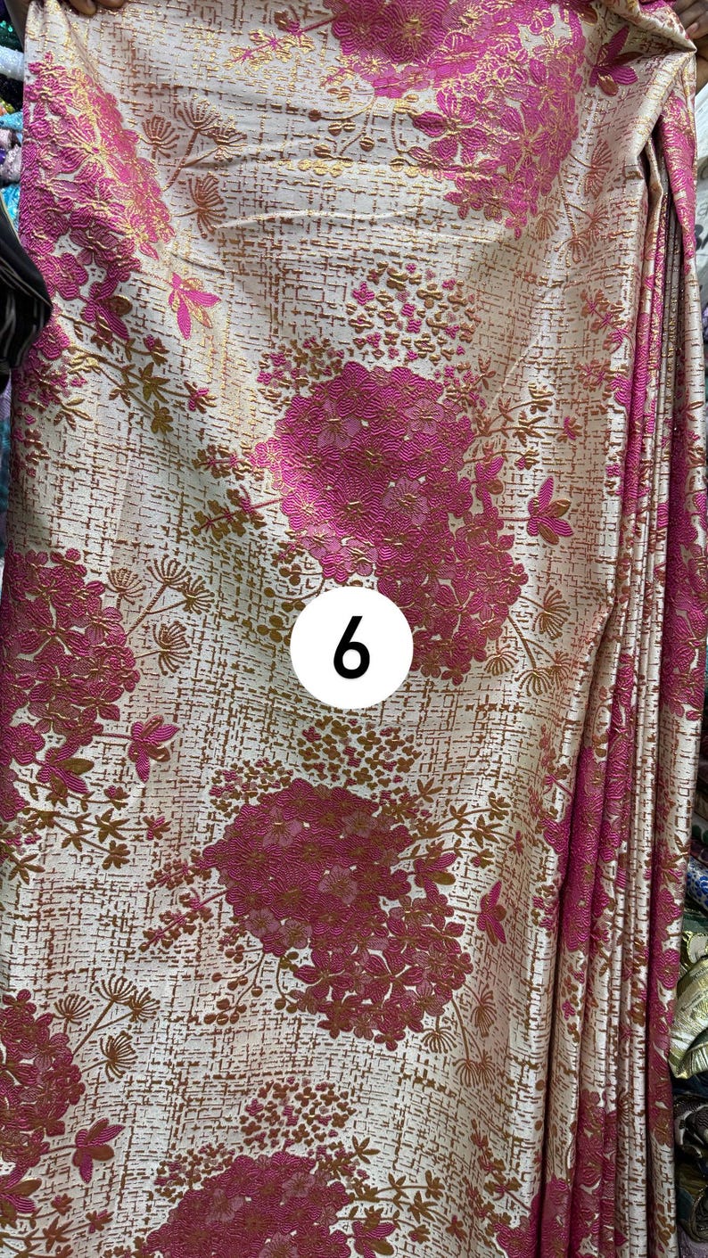 May include: A fabric featuring a cream background with a textured, metallic gold grid pattern and large, vibrant magenta floral clusters. A white circular sticker with the black number 6 is positioned in the center of the textile design.
