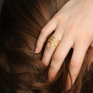 May include: A gold ring with a delicate, leafy design. The ring is worn on the index finger of a hand with brown hair.