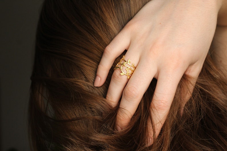 May include: A gold ring with a delicate, leafy design. The ring is worn on the index finger of a hand with brown hair.