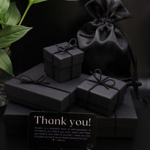 May include: Three black gift boxes with black ribbon bows, stacked on top of each other. A black satin pouch is in the background. A small card with the text "Thank you! Jewelry is a beautiful form of self-expression. It introduces you before you even speak and helps you explore new sides of yourself. I hope you love this piece as much as we loved making it for you! - Katrina" is in the foreground.