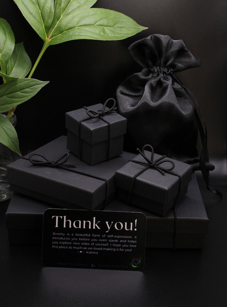 May include: Three black gift boxes with black ribbon bows, stacked on top of each other. A black satin pouch is in the background. A small card with the text "Thank you! Jewelry is a beautiful form of self-expression. It introduces you before you even speak and helps you explore new sides of yourself. I hope you love this piece as much as we loved making it for you! - Katrina" is in the foreground.