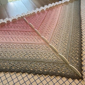 Op de afbeelding: Een driehoekige gehaakte sjaal met een kleurverloop van lichtroze naar donkergrijs. De sjaal is vastgezet op een blokkeermat met witte ronde blokkeerspelletjes langs de randen, wat een ingewikkeld opengewerkt kantpatroon laat zien.