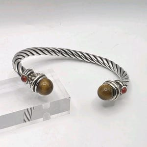 May include: A silver-toned cuff bracelet featuring a twisted cable design. The open ends are capped with large, spherical brown tiger's eye gemstones, flanked by smaller, round, reddish-orange accent stones set in decorative metal mounts.