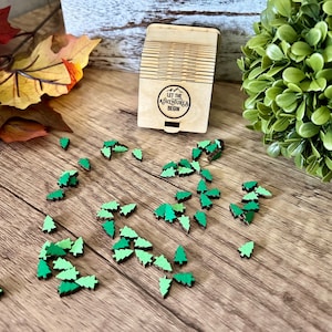May include: A wooden box with the text "Let the Adventures Begin" on the front. Scattered around the box are small wooden green pine tree ornaments.