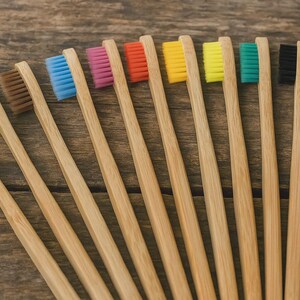 Könnte beinhalten: Eine Ansammlung von Bambus-Zahnbürsten mit unterschiedlich farbigen Borsten, darunter weiß, grün, braun, blau, pink, rot, gelb und schwarz. Die Zahnbürsten sind auf einer Holzoberfläche angeordnet und zeigen ihre natürlichen Bambusgriffe.