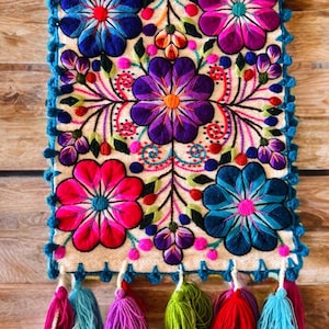 May include: Embroidered textile with a cream-colored background, featuring large, colorful flowers in shades of blue, pink, and purple. The edges are trimmed with blue crochet, and the bottom has tassels in various colors. A vibrant, decorative piece.