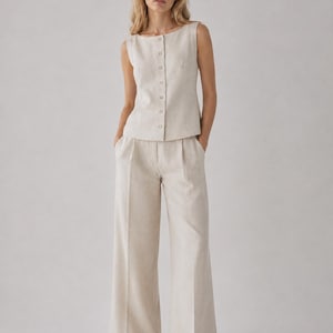 May include: A sleeveless beige top with a vertical row of buttons down the front, paired with matching wide-leg trousers featuring a pleated waist and a straight-leg silhouette. The set is crafted from a textured, light-colored fabric.