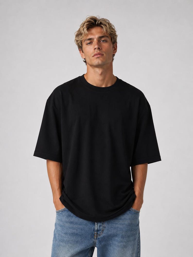 May include: A black, oversized t-shirt with short sleeves. The shirt is made of a soft material and is being worn with blue jeans. The model is standing in front of a plain, light gray background.