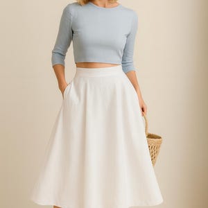 Puede incluir: Un top corto azul claro de manga tres cuartos combinado con una falda midi blanca. La falda tiene cintura alta y bolsillos. Sandalias de cuero marr&oacute;n y una cesta de mimbre completan el conjunto.