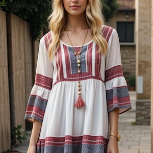 Puede incluir: Un vestido blanco de manga corta con rayas bordadas rojas y azules. El vestido tiene un escote redondo y una falda fruncida. La modelo lleva un collar de oro de varias capas con un borla.