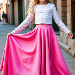 May include: A young girl wearing a white long-sleeved crop top and a bright pink full-length skirt. She is standing on a brick walkway.