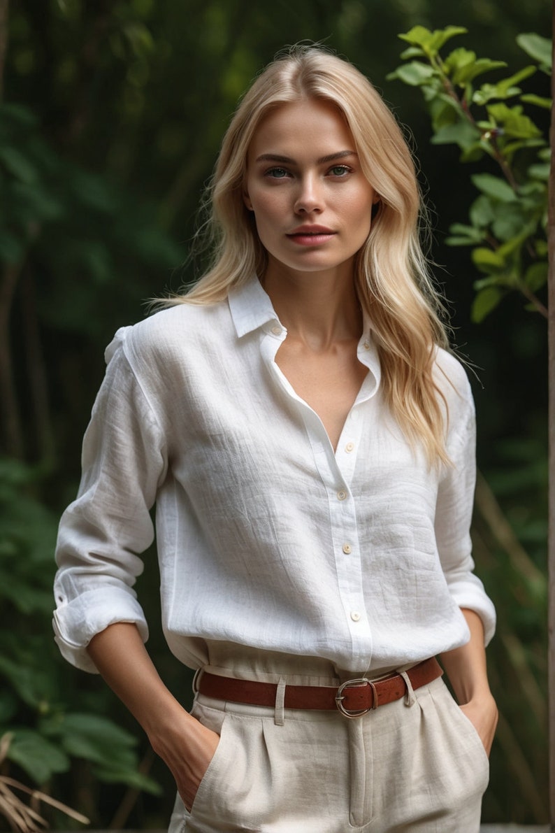 Puede incluir: Una mujer con una camisa de lino blanca con un cintur&oacute;n de cuero marr&oacute;n y pantalones de lino beige. Est&aacute; de pie en un jard&iacute;n con follaje verde en el fondo.