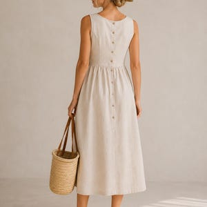 May include: A sleeveless beige midi dress featuring a vertical row of buttons down the back, paired with a straw hat with a brown band and a woven straw bucket bag. The dress has a fitted bodice and a gathered skirt, worn with brown sandals.