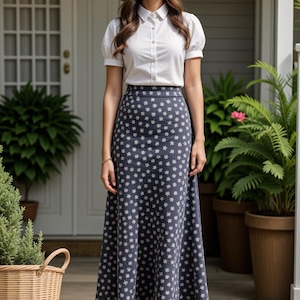 May include: A woman wearing a white button-down shirt with a blue and white floral print skirt and a straw hat. She is standing in front of a house with a wooden door.