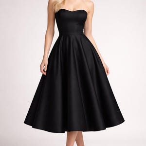 May include: A strapless, knee-length black dress with a sweetheart neckline. The dress has a fitted bodice and a full, flared skirt. The model is wearing black high heels. The background is plain white.