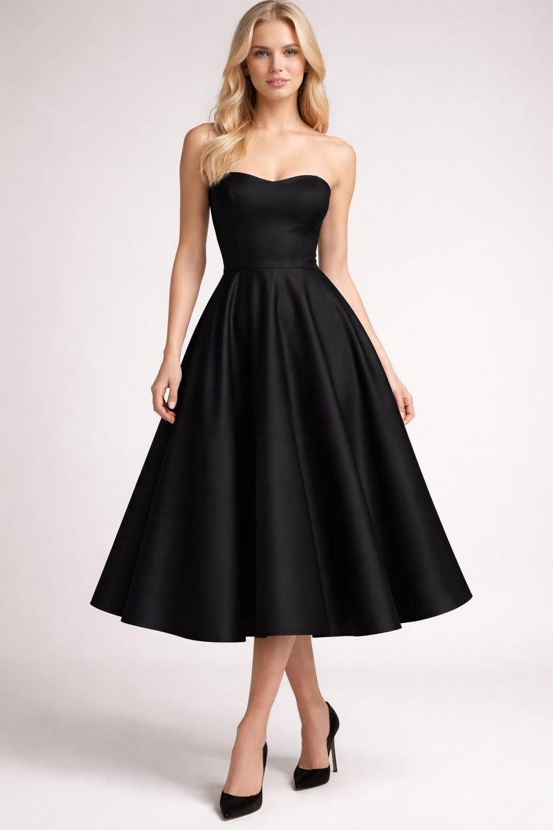 May include: A strapless, knee-length black dress with a sweetheart neckline. The dress has a fitted bodice and a full, flared skirt. The model is wearing black high heels. The background is plain white.