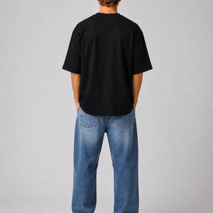 May include: A person wearing a black short-sleeved t-shirt and blue denim jeans. The jeans have a relaxed fit. The person is also wearing white sneakers. The background is a solid light gray.