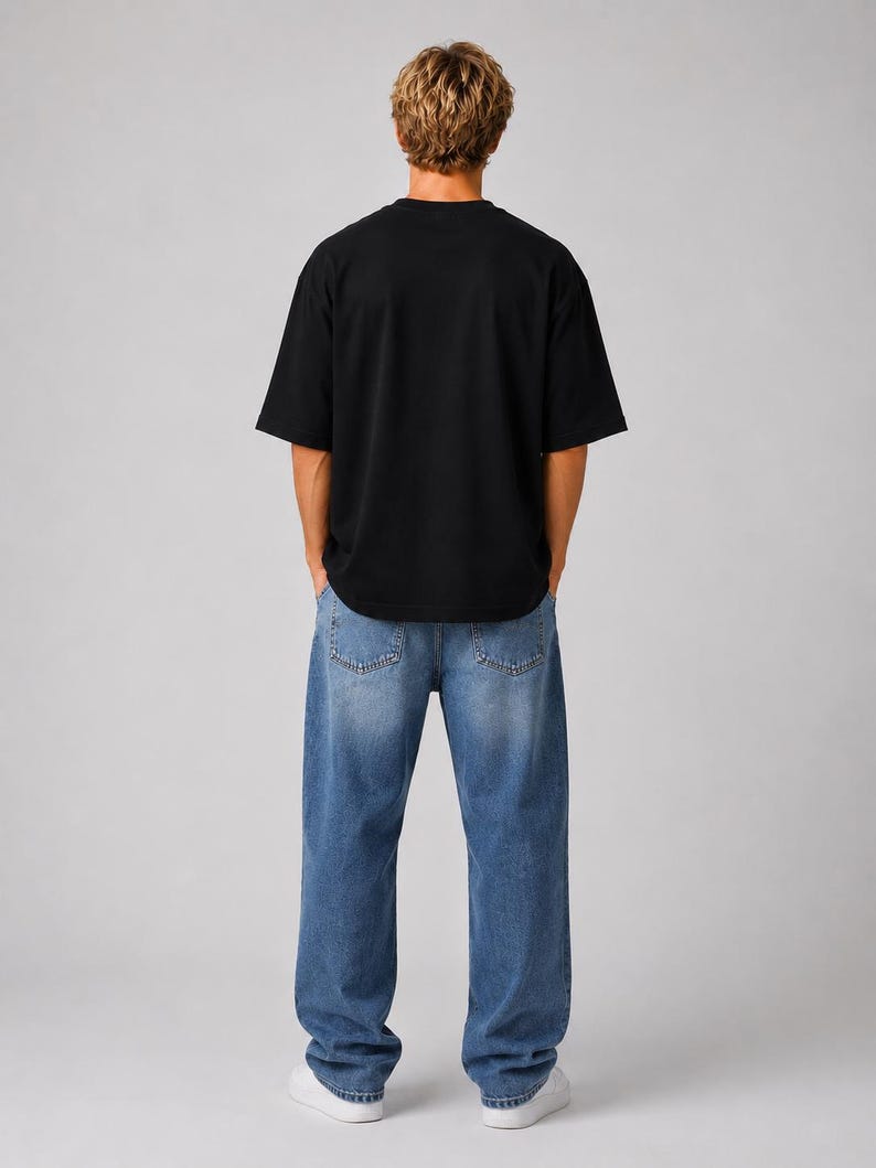 May include: A person wearing a black short-sleeved t-shirt and blue denim jeans. The jeans have a relaxed fit. The person is also wearing white sneakers. The background is a solid light gray.