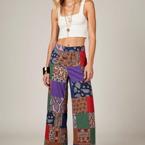 May include: A woman wearing a white crop top and wide-leg patchwork pants with a variety of colors and patterns. She is wearing gold jewelry and brown sandals.