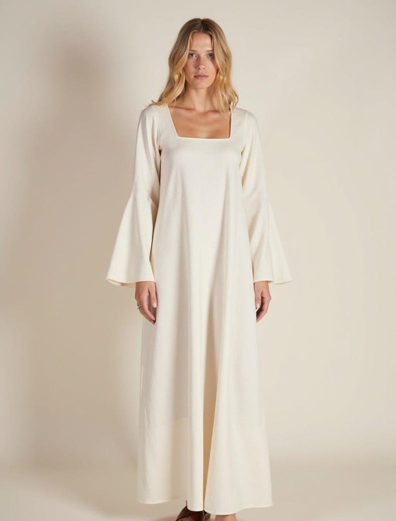 May include: A long, white maxi dress with a square neckline and long, wide bell sleeves. The dress is made from a lightweight fabric and has a flowing silhouette.