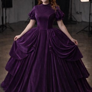 Puede incluir: Un vestido largo de terciopelo morado con cuello alto y mangas cortas abullonadas. El vestido presenta una falda en capas y lo lleva una persona que lleva una corona de joyas. El fondo es un estudio con equipo de iluminaci&oacute;n.