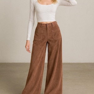 May include: A woman wearing a white long-sleeve scoop neck crop top and brown corduroy wide-leg pants. The pants have two front pockets and are paired with white sneakers.