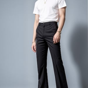 May include: A man wearing a white t-shirt and black flared pants. He is wearing a gold chain necklace and a watch on his left wrist. He is standing on a gray background.