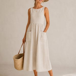 May include: A sleeveless midi dress in a light beige color featuring a round neckline, a fitted bodice, and a gathered skirt with two large front patch pockets. The outfit includes a wide-brimmed straw hat, tan leather sandals, and a woven straw tote bag.