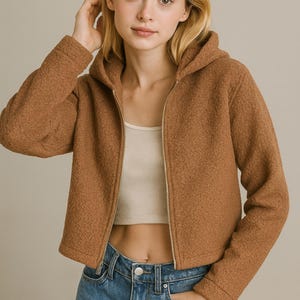 May include: A brown hooded jacket with a full-length zipper. The jacket has a textured, fuzzy appearance and is paired with a white crop top and blue jeans. The model is wearing the jacket.