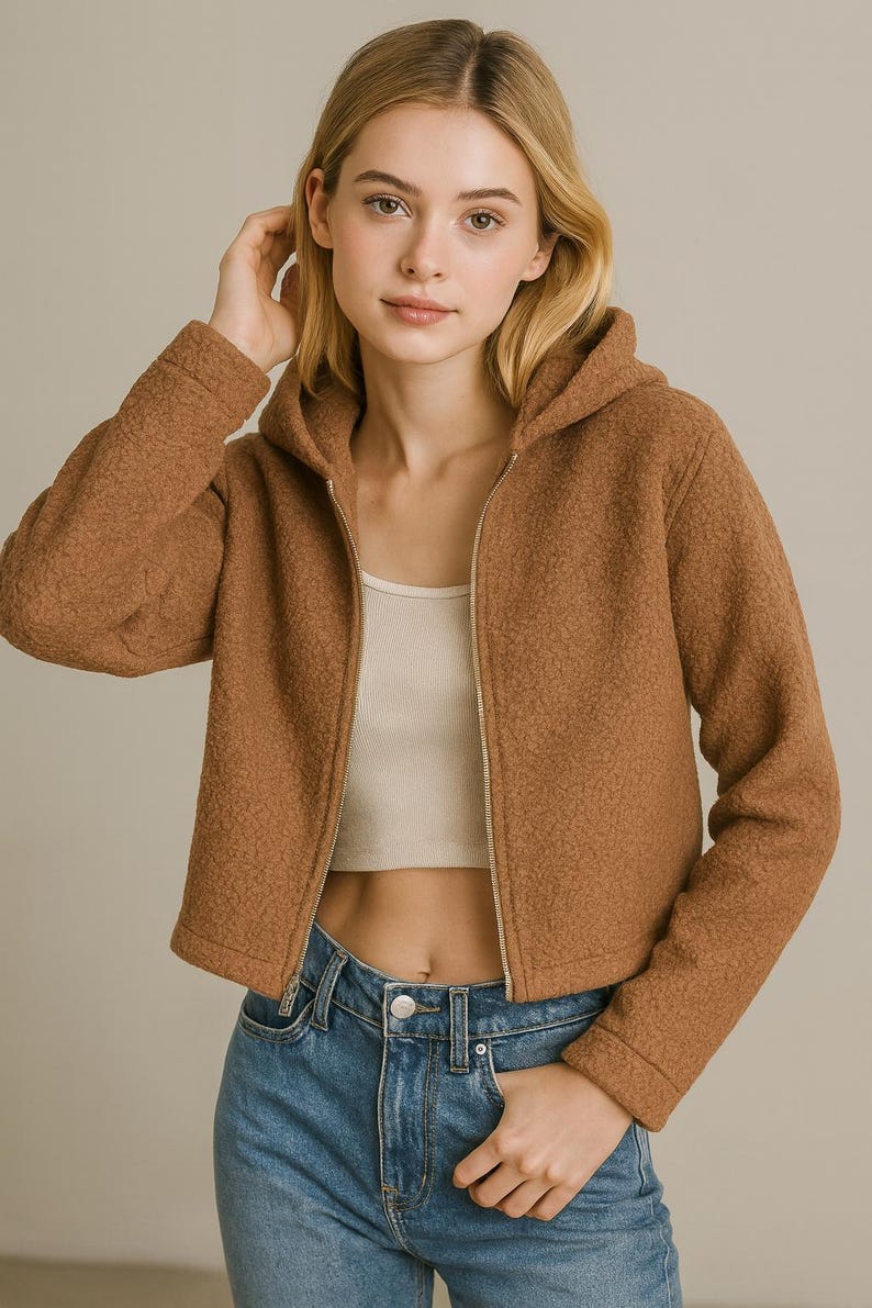 May include: A brown hooded jacket with a full-length zipper. The jacket has a textured, fuzzy appearance and is paired with a white crop top and blue jeans. The model is wearing the jacket.