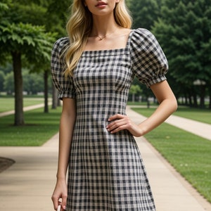 May include: A woman wearing a black and white gingham dress with puffed sleeves. The dress has a square neckline and a fitted bodice. She is holding a small, light-colored handbag.