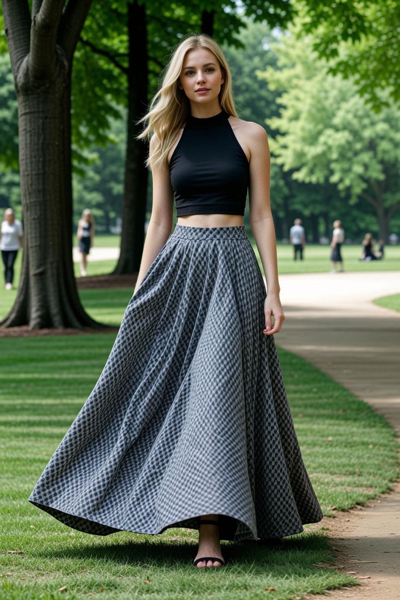 May include: A woman wearing a black halter crop top and a long, flowing skirt with a gray and white checkered pattern. The skirt is full and reaches the ground. The woman is walking on a path in a park.