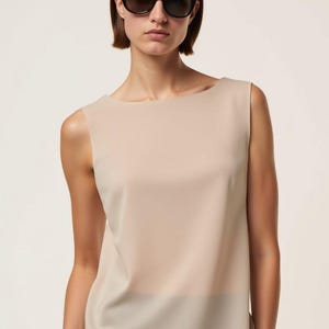 May include: A woman wearing a beige sleeveless top with a scoop neck and black sunglasses.