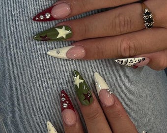 May include: A close-up of a hand with long, pointed acrylic nails. The nails feature various designs, including white, red, and olive green colors. Some nails have star and gem accents, while others have a French tip design.