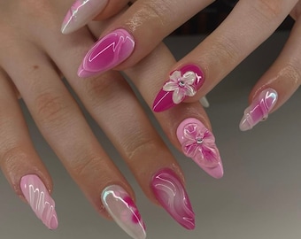 May include: A set of long, pointed acrylic nails in shades of pink and white. Some nails feature 3D floral embellishments, while others have swirled or marbled designs. The nails are well-manicured and showcase a variety of textures and finishes.
