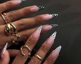 May include: Long, pointed fingernails with a white and black polka dot design. The nails have a natural pink base with a white tip and black polka dots. Gold rings adorn the fingers.