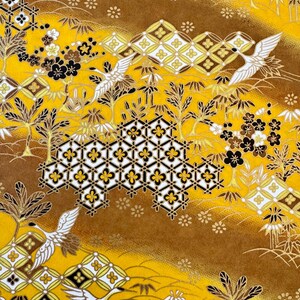 May include: A yellow fabric with a repeating pattern of white cranes, black and white flowers, and a geometric design. The fabric has a brown background with a subtle gradient.