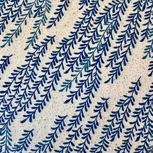 May include: A close-up of a white fabric featuring a repeating pattern of dark blue and teal fern-like branches. The branches are arranged diagonally across the fabric, creating a textured design. The background has a speckled appearance, adding visual interest.