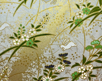 May include: A white background with a gold and silver pattern of flowers, leaves, and rabbits. The flowers are white with green centres and blue accents. The leaves are green and the rabbits are white.