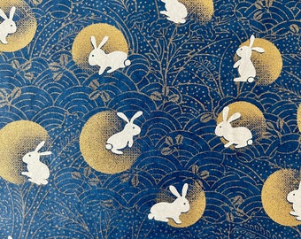 May include: A blue and gold patterned wrapping paper with white rabbits and moons. The rabbits are in various poses, some are sitting, some are standing, and some are hopping. The moons are full and have a gold outline.