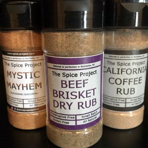 May include: Three glass jars of spice blends with black lids. The labels are purple and white with the text "The Spice Project" and the names of the blends: "Mystic Mayhem", "Beef Brisket Dry Rub", and "California Coffee Rub". Each label also includes the text "Small Batch", "All Natural", "Preservative Free", and "Gluten Free".