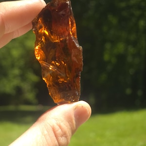 May include: A piece of amber-coloured glass with a jagged, uneven form. The light shines through the glass, creating a bright effect. The background is a blurred green, suggesting an outdoor setting.