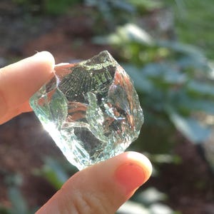 May include: A clear, faceted glass rock with a light green tint is held up to the sunlight. The edges of the glass are sharp and reflect the light, creating a sparkling effect. The background is blurred with green foliage.