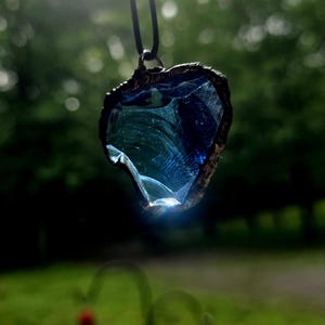 May include: A heart-shaped pendant with a dark blue, textured glass centre and a bronze-coloured metal frame. The pendant hangs from a black cord, with a blurred green background.