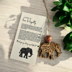 Puede incluir: Un adorno de elefante de madera con tallas intrincadas y la palabra "hermandad" inscrita en &eacute;l. El adorno cuelga de una cuerda y tiene una peque&ntilde;a cuenta marr&oacute;n. El adorno se acompa&ntilde;a de una tarjeta con el texto "A story of love & loyalty" y una silueta negra de un elefante.