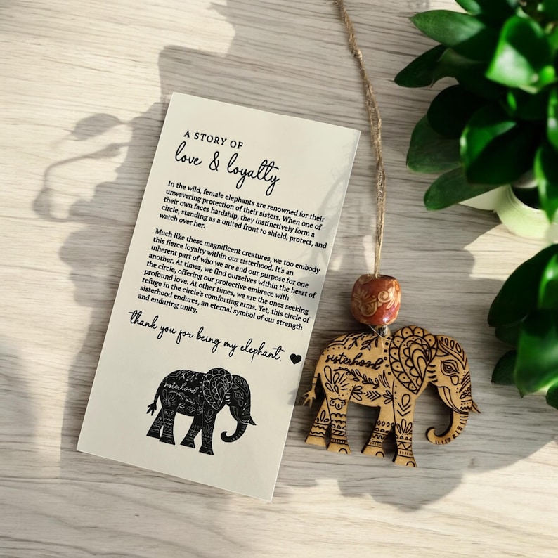 Puede incluir: Un adorno de elefante de madera con tallas intrincadas y la palabra "hermandad" inscrita en &eacute;l. El adorno cuelga de una cuerda y tiene una peque&ntilde;a cuenta marr&oacute;n. El adorno se acompa&ntilde;a de una tarjeta con el texto "A story of love & loyalty" y una silueta negra de un elefante.