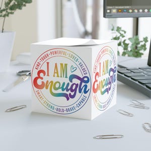 Op de afbeelding: Een witte kubusvormige notitieblok met de tekst "I AM Enough" in een regenboogontwerp. De tekst is omgeven door een cirkel met positieve affirmaties. Er liggen paperclips verspreid.