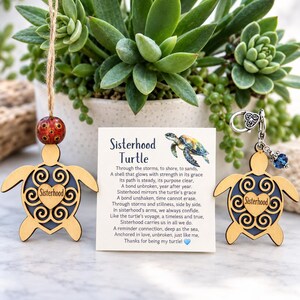 May include: Two wooden turtle ornaments featuring a cutout heart design with the word Sisterhood. One turtle hangs from a jute string with a red patterned bead, while the other has a silver metal lobster clasp and a small blue crystal bead.