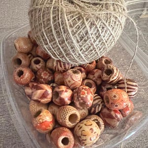 Puede incluir: Un recipiente de pl&aacute;stico transparente lleno de cuentas de madera de varios tipos. Las cuentas son marrones y naranjas con intrincados patrones. Una bola de hilo est&aacute; encima de las cuentas.