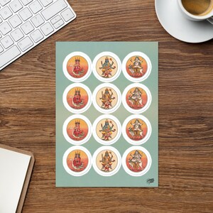 Op de afbeelding: Een vel stickers met kleurrijke illustraties van hindoe-goden in witte cirkels. Elke afbeelding heeft een levendige oranje en gele achtergrond. De stickers zijn gerangschikt op een lichtgroene achtergrond.